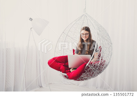 Beautiful woman in eyeglasses in fashionable armchair with laptop. Girl with pc in modern interior at red cushions. Stylish portrait of freelancer, work at home concept 135501772