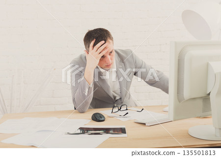 Working problems, issues of business. Young businessman in white modern office interior, thoughtful, sitting at computer table. Working problems, issues of business. Young businessman in white modern office interior, thoughtful, sitting at computer table. 135501813