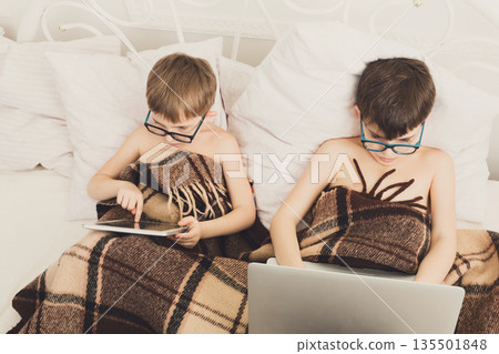 Boys in parents' bed at morning with laptop and tablet. Brothers play computer games. Siblings and gadgets. Children in glasses. Boys in parents' bed at morning with laptop and tablet. Brothers play computer games. Siblings and gadgets. Children in glasses. 135501848