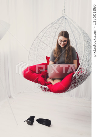 Beautiful woman in eyeglasses in fashionable armchair with laptop. Girl with pc in modern interior at red cushions. Stylish portrait of freelancer, work at home concept Beautiful woman in eyeglasses in fashionable armchair with laptop. Girl with pc in modern interior at red cushions. Stylish portrait of freelancer, work at home concept 135501860