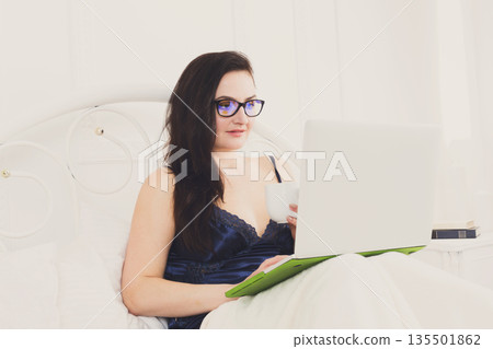 Sunday morning in bed. Middle aged woman in blue nightwear surfing internet at her laptop computer, copy space Sunday morning in bed. Middle aged woman in blue nightwear surfing internet at her laptop computer, copy space 135501862