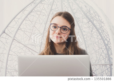 Beautiful smiling woman in eyeglasses in fashionable armchair with laptop. Girl with pc in modern interior. Stylish portrait of freelancer, work at home concept Beautiful smiling woman in eyeglasses in fashionable armchair with laptop. Girl with pc in modern interior. Stylish portrait of freelancer, work at home concept 135501866