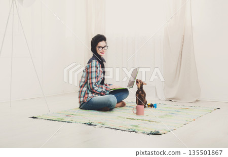 Young girl in computer glasses on carpet with laptop and dog chihuahua. Hipster freelancer woman works at home. Home office, freelance. Young girl in computer glasses on carpet with laptop and dog chihuahua. Hipster freelancer woman works at home. Home office, freelance. 135501867
