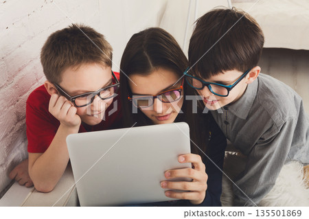 Group of kids in eye glasses look into tablet. Children computer games, social networks and media addiction concept. Girl and boys with gadget. Communication technologies. 135501869