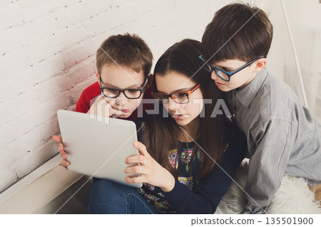 Group of kids in eye glasses look into tablet. Children computer games, social networks and media addiction concept. Girl and boys with gadget. Communication technologies. 135501900