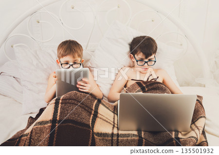 Boys in parents' bed at morning with laptop and tablet. Brothers play computer games. Siblings and gadgets. Children in glasses. 135501932