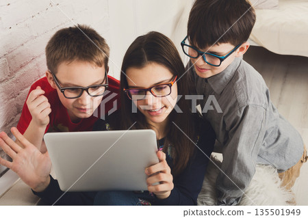 Group of kids in eye glasses look into tablet. Children computer games, social networks and media addiction concept. Girl and boys with gadget. Communication technologies. 135501949
