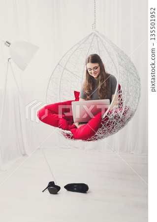 Beautiful woman in eyeglasses in fashionable armchair with laptop. Girl with pc in modern interior at red cushions. Stylish portrait of freelancer, work at home concept Beautiful woman in eyeglasses in fashionable armchair with laptop. Girl with pc in modern interior at red cushions. Stylish portrait of freelancer, work at home concept 135501952