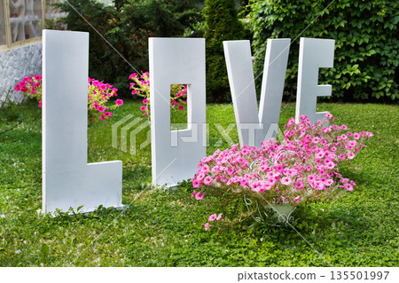 Love word outdoors. Big white plastic letters on the grass near flowers in the garden, wedding ceremony decoration, romantic holiday decor. 135501997