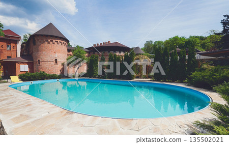 Luxury swimming pool beside the hotel in medieval british castle style. Landscape design of resort territory. Blue water in summer park. Luxury swimming pool beside the hotel in medieval british castle style. Landscape design of resort territory. Blue water in summer park. 135502021