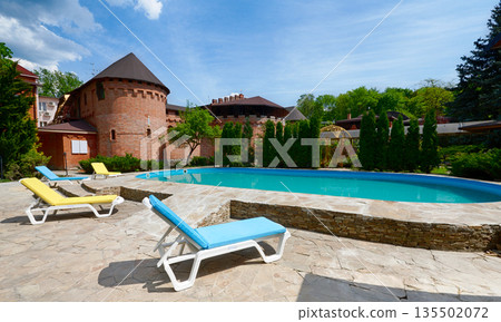 Luxury swimming pool beside the hotel in medieval british castle style. Landscape design of resort territory. Swimming pool outdoors. Blue water in summer park. Luxury swimming pool beside the hotel in medieval british castle style. Landscape design of resort territory. Swimming pool outdoors. Blue water in summer park. 135502072