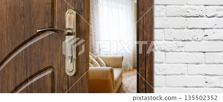 Half opened door handle closeup, entrance to a living room. View of sofa. Welcome, privacy concept. Door lock with keys, white brick wall, modern interior design. Half opened door handle closeup, entrance to a living room. View of sofa. Welcome, privacy concept. Door lock with keys, white brick wall, modern interior design. 135502352