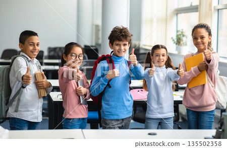 Education Concept. Diverse group of happy smiling international classmates standing in line with notebooks and backpacks in classroom and showing thumbs up sign gesture, enjoying studying at school Education Concept. Diverse group of happy smiling international classmates standing in line with notebooks and backpacks in classroom and showing thumbs up sign gesture, enjoying studying at school 135502358