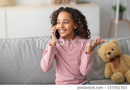 Communication concept. Happy african american girl talking on smartphone, enjoying conversation with friend. Teen sitting on sofa with teddy bear nearby Communication concept. Happy african american girl talking on smartphone, enjoying conversation with friend. Teen sitting on sofa with teddy bear nearby 135502383