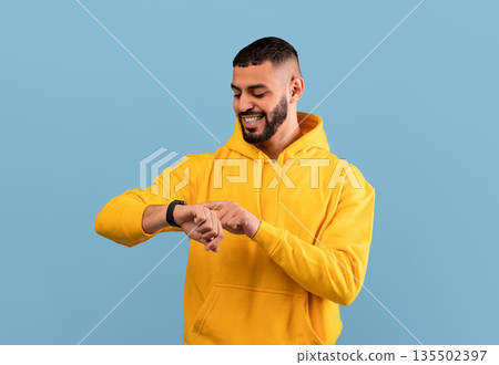 Young arab guy checking his smartwatch on blue studio background. Happy middle eastern man using fitness tracker. Modern technologies and active lifestyle concept Young arab guy checking his smartwatch on blue studio background. Happy middle eastern man using fitness tracker. Modern technologies and active lifestyle concept 135502397