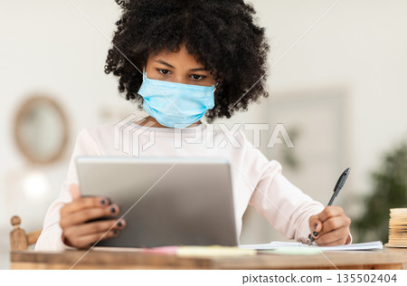 African Girl With Tablet Learning Online Wearing Mask Sitting At Desk At Home. Virus Protection At School. Selective Focus 135502404