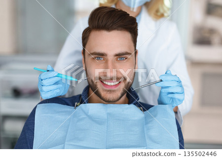 Smiling young bearded man sitting in dental chair, visiting modern dental clinic, having regular checkup, unrecognizable woman dentist holding dental tools next to male patient mouth 135502446