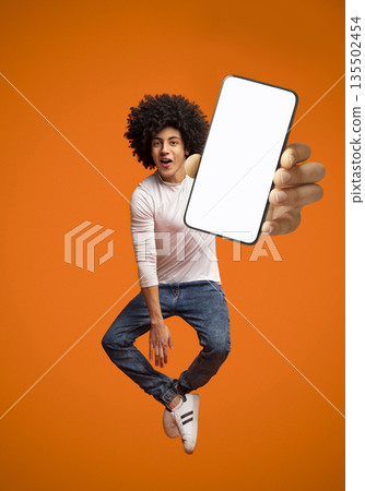 Funny young black teen guy jumping on air, showing cellphone with white empty screen on orange studio background. Young man demonstrating mockup copy space for your mobile app design 135502454