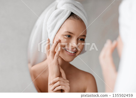 Portrait of young lady applying nourishing eye cream after shower, looking at mirror and smiling in bathroom interior. Anti-aging cosmetics, tools against lines, wrinkles eye care concept 135502459