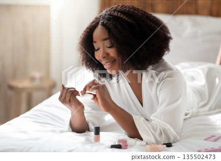 Beautiful African American woman in bathrobe doing her nails on bed at home, copy space. Lovely black lady making manicure, pampering herself, having domestic spa day 135502473