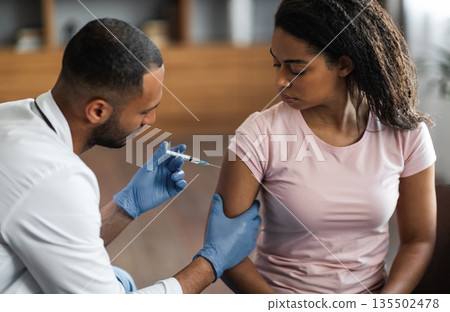 Handsome african american man making injection in shoulder for female patient, wearing blue gloves. Young black lady getting vaccinated against coronavirus at clinic, closeup shot 135502478