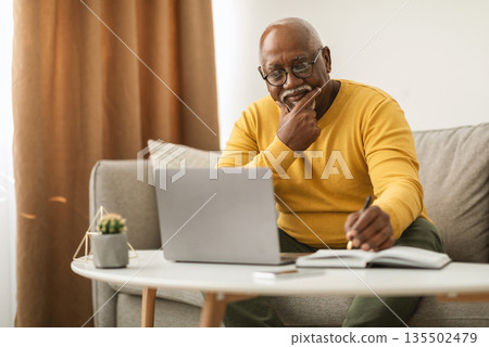 Mature African American Business Man Using Laptop And Writing Taking Notes Sitting On Couch Indoor. Senior Male Freelancer Working Online On Computer At Home. E-Learning In Older Age Mature African American Business Man Using Laptop And Writing Taking Notes Sitting On Couch Indoor. Senior Male Freelancer Working Online On Computer At Home. E-Learning In Older Age 135502479