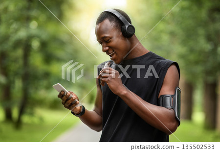Cheerful african sportsman eating protein bar and using smartphone while resting, exercising at park, empty space Cheerful african sportsman eating protein bar and using smartphone while resting, exercising at park, empty space 135502533