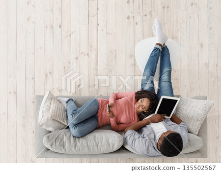 Weekend Leisure. Young Black Couple Relaxing On Couch With Digital Tablet And Smartphone, Top View With Copy Space 135502567