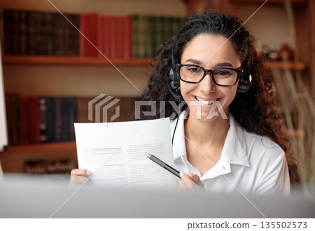 Distant Remote Work. Portrait of smiling young woman in eyeglasses and headset having virtual videocall using laptop, holding paper and showing report, contract document or agreement to web camera Distant Remote Work. Portrait of smiling young woman in eyeglasses and headset having virtual videocall using laptop, holding paper and showing report, contract document or agreement to web camera 135502573