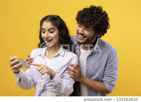 Excited indian couple looking at smartphone screen, using new app or reading cool news, standing over yellow studio background. Happy man and woman using cellphone, guy embracing girlfriend 135502610
