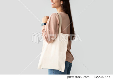 Close Up Of Young Female Consumer Holding Blank White Eco Tote Bag, Posing Standing Over Studio Background. Lady Carrying Flax Tote Shopper Handbag. Eco-Friendly Lifestyle, Stop Using Plastic Concept Close Up Of Young Female Consumer Holding Blank White Eco Tote Bag, Posing Standing Over Studio Background. Lady Carrying Flax Tote Shopper Handbag. Eco-Friendly Lifestyle, Stop Using Plastic Concept 135502633