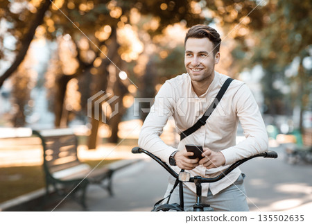 Street style. Young stylish man texting on phone while sitting on bike in park, free space 135502635