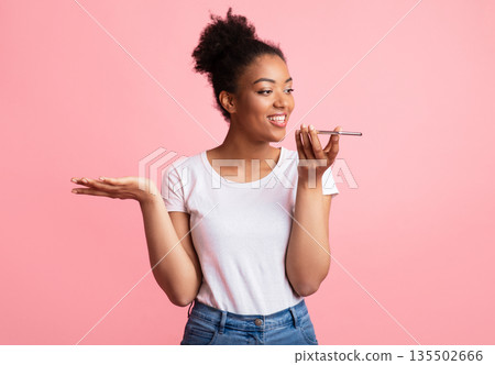 Happy young black lady using voice assistant on smartphone, gesturing isolated over pastel pink studio wall, copyspace Happy young black lady using voice assistant on smartphone, gesturing isolated over pastel pink studio wall, copyspace 135502666