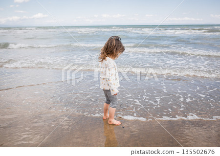 side view of little girl with brown hair standing on sand along the seashore side view of little girl with brown hair standing on sand along the seashore 135502676