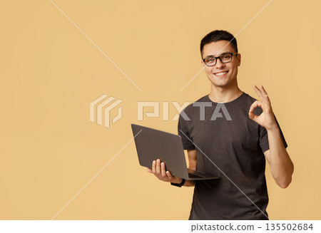 Excellent result, good feedback and win. Smiling young handsome male in glasses expresses emotions of success shows okay gesture and holds laptop, isolated on light background free space, studio shot Excellent result, good feedback and win. Smiling young handsome male in glasses expresses emotions of success shows okay gesture and holds laptop, isolated on light background free space, studio shot 135502684