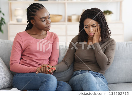 African american young lady comforting her ill girlfriend or sister, sitting on sofa at home, copy space. Black woman suffering from toothache, touching her cheek, feeling sick African american young lady comforting her ill girlfriend or sister, sitting on sofa at home, copy space. Black woman suffering from toothache, touching her cheek, feeling sick 135502715