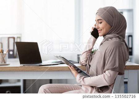Muslim business lady in hijab holding clipboard, checking documentation and talking on mobile phone with partner while sitting at workplace in office 135502716