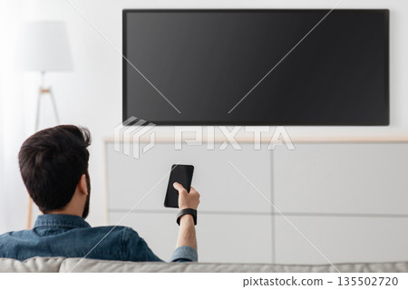 Young man watching television with blank screen and using smart TV remote control application on smartphone, back view. Select movie stream, change channel or settings in the menu 135502720