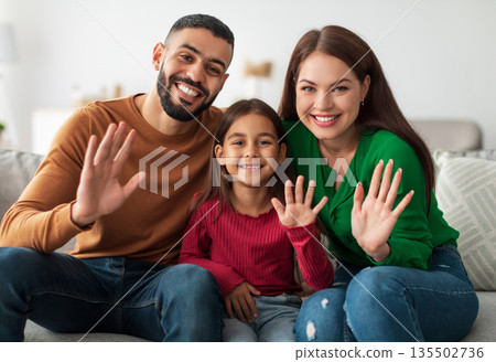 Hello. Cheerful Young Middle Eastern Family Of Three People Waving Hand Smiling To Camera Sitting On Couch At Home. Hi Greeting Gesture Concept. Video Call And Modern Remote Communication 135502736