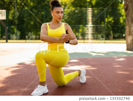 Sport And Healthy Lifestyle. Fit black woman doing curtsy lunges with twist, exercising outdoors on basketball stadium. Portrait of confident lady training in wireless earbuds, full body length Sport And Healthy Lifestyle. Fit black woman doing curtsy lunges with twist, exercising outdoors on basketball stadium. Portrait of confident lady training in wireless earbuds, full body length 135502740