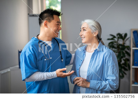 Cheerful millennial japanese man doctor meets and talks with aged female patient at exam in clinic office interior. Meeting in hospital, treatment with therapist, medical health care and good service 135502756