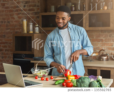 Happy african-american man preparing vegetable salad while looking on laptop Happy african-american man preparing vegetable salad while looking on laptop 135502765