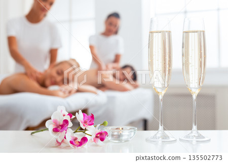 Romantic Spa Vacations. Relaxed Couple Enjoying Back Massage Lying On Beds At Beauty Center Indoor. Focus On Tropic Orchid Flowers And Champlagne Glasses. Honeymoon Relaxation. Selective Focus 135502773