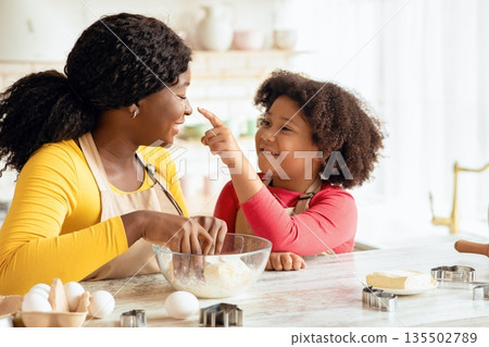 Cute Little Black Daughter And Her Mom Having Fun While Cooking Together In Kitchen, Cheerful African Family Preparing Homemade Pastry And Fooling, Funny Little Girl Playfully Touching Mother's Nose Cute Little Black Daughter And Her Mom Having Fun While Cooking Together In Kitchen, Cheerful African Family Preparing Homemade Pastry And Fooling, Funny Little Girl Playfully Touching Mother's Nose 135502789