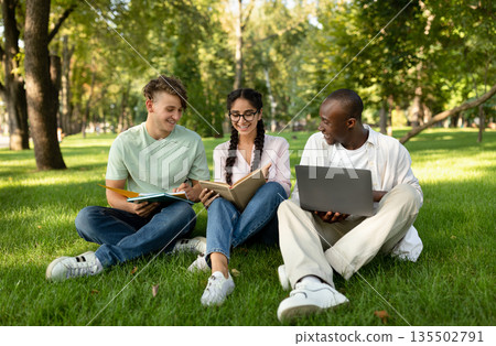 Group of diverse students friends learning, reading books, using laptop and discussing something in university campus park, sitting on grass outside 135502791