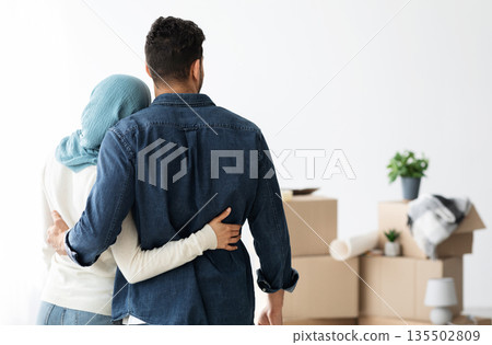 Back view of young muslim family husband and wife cuddling while moving to new house, copy space. Arab man and lady in hijab standing in empty white room, looking at paper boxes and hugging Back view of young muslim family husband and wife cuddling while moving to new house, copy space. Arab man and lady in hijab standing in empty white room, looking at paper boxes and hugging 135502809
