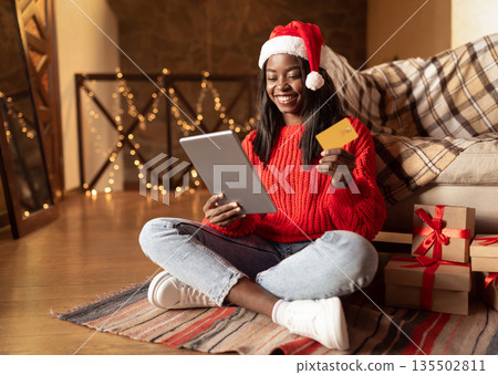 Online holiday sale. Happy black lady in Santa hat using digital tablet and credit card to buy Christmas gifts on web from home, copy space. Young woman buying presents on internet via touch pad 135502811