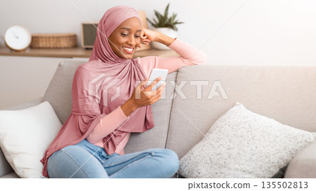 Portrait of millennial Muslim lady in hijab using mobile phone at home, messaging or browsing social media, sitting on couch in living room. Pretty Arab woman checking email, talking to friend online Portrait of millennial Muslim lady in hijab using mobile phone at home, messaging or browsing social media, sitting on couch in living room. Pretty Arab woman checking email, talking to friend online 135502813