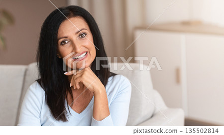 Aged Beauty. Portrait of relaxed mature woman with long dark hair sitting on the couch in living room and posing. Tranquil positive lady looking and smiling at the camera, selective focus 135502814