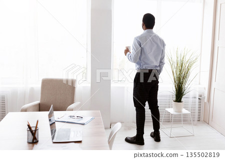 Unrecognizable african american businessman or manager drinking coffee near panoramic window in office, having break after successful deal, copy space, back view, full length shot Unrecognizable african american businessman or manager drinking coffee near panoramic window in office, having break after successful deal, copy space, back view, full length shot 135502819